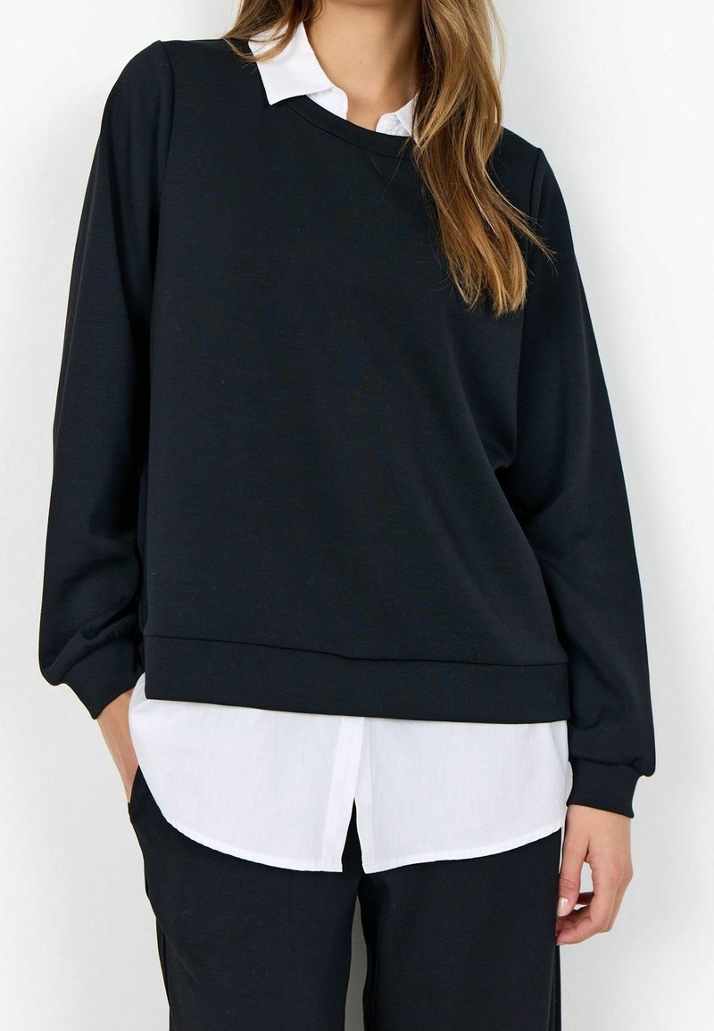 Femme portant un pull-over noir à manches longues sur une chemise blanche à boutons, les mains partiellement dans les poches d'un pantalon noir.