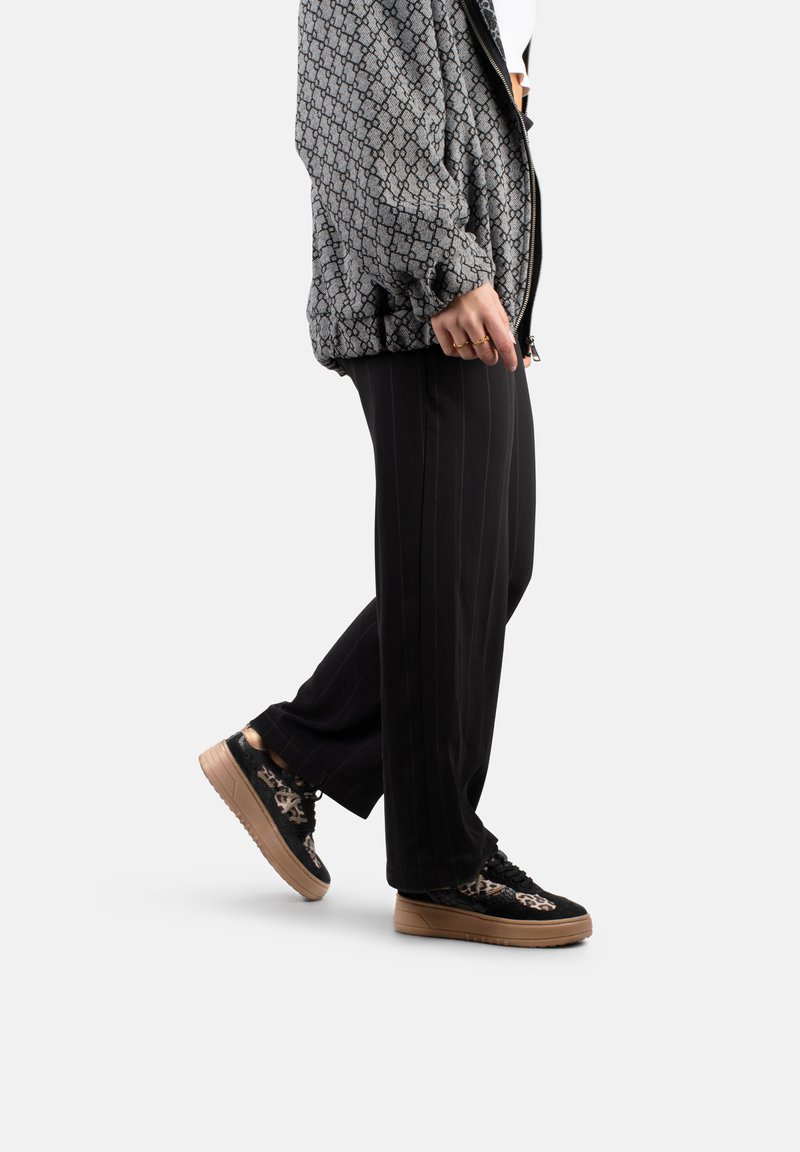 Bomber jacket a motivi neri e grigi sopra una maglietta bianca, abbinato a pantaloni neri a righe e sneakers nere con suole e dettagli beige.