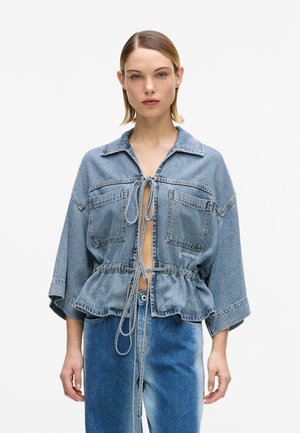 Femme portant une veste en jean courte bleu clair avec des cordons à nouer, de grandes poches avant et des manches larges, associée à un jean bicolore bleu.