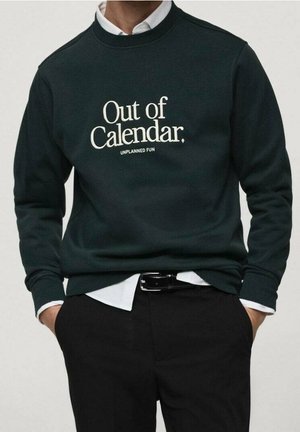 Osoba nosząca ciemnozieloną bluzę z białym napisem "Out of Calendar. UNPLANNED FUN" na białej koszuli z kołnierzykiem i czarnych spodniach.