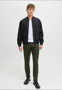 Black bomber jacket over a white t-shirt, paired with olive green slim-fit pants and black lace-up shoes, standing against a neutral background.