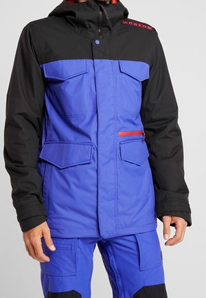 Mann trägt eine blau-schwarze Jacke mit mehreren Taschen, einem roten Reißverschlussdetail und passende blaue Hosen mit Cargotaschen.