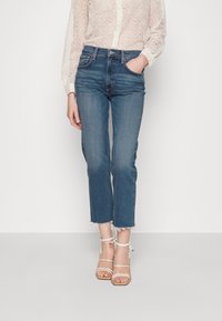 Jeans cortos de color azul claro con dobladillo deshilachado, combinados con una blusa de encaje color crema. Lleva sandalias de tiras blancas con suela de color beige.