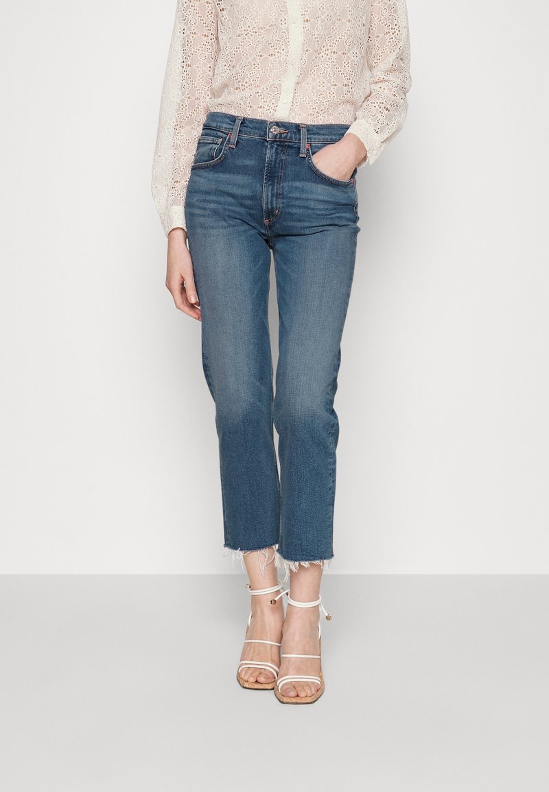 Jeans cortos de color azul claro con dobladillo deshilachado, combinados con una blusa de encaje color crema. Lleva sandalias de tiras blancas con suela de color beige.