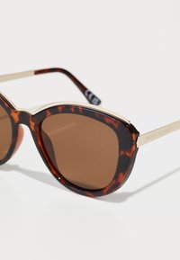 Óculos de sol tartaruga com lentes castanhas, detalhes em metal dourado e formato de olho de gato. Apresentam um acabamento liso e brilhante na armação.