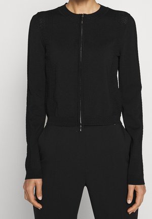 Femme portant un pull noir cintré à manches longues zippé avec des détails en tricot texturé et un pantalon noir, les mains détendues le long du corps.