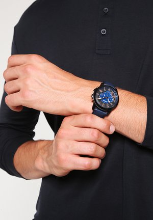 Person iført sort langærmet skjorte, der justerer et blå læderrem armbåndsur med sort og blå urskive med romertal.