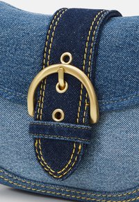 Gros plan sur un sac en denim bleu avec une sangle bleu foncé, des surpiqûres jaunes et une boucle en métal doré fixée à travers des œillets.