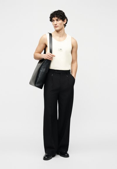 Νέος άνδρας που φοράει λευκό αμάνικο πουκάμισο και μαύρο παντελόνι με φαρδιά πόδια, κρατώντας μια μεγάλη μαύρη δερμάτινη τσάντα ώμου πάνω στον έναν ώμο.