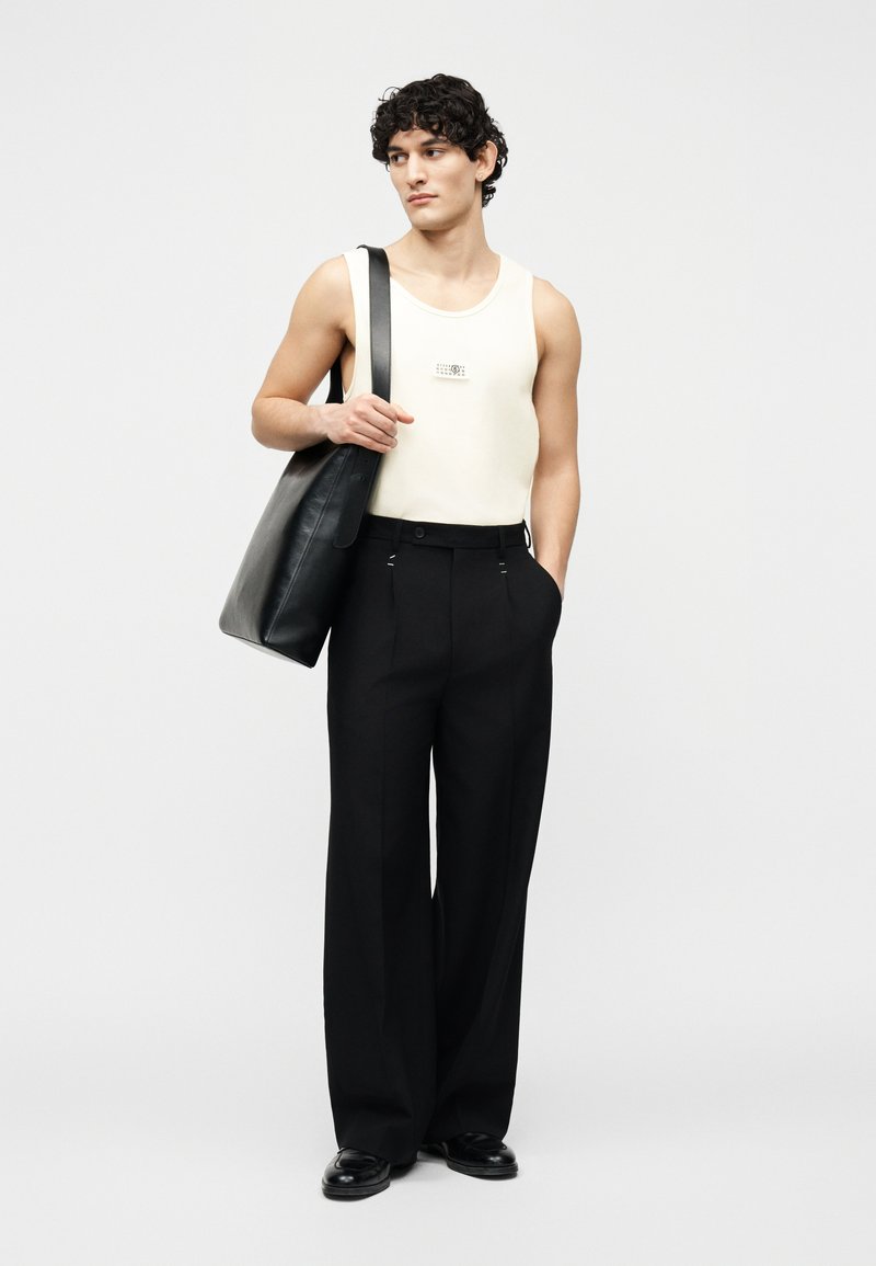 Giovane uomo con indosso una canottiera bianca e pantaloni neri a gamba larga, che tiene una grande borsa tote in pelle nera su una spalla.