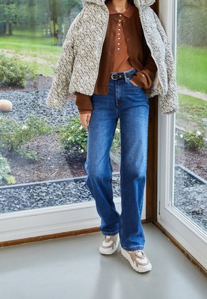 Person trägt eine gemusterte beige Daunenjacke über einem braunen, mit Knöpfen versehenen Pullover, blaue Jeans mit schwarzem Gürtel und beige-weiße Turnschuhe, steht drinnen neben einer Glastür.