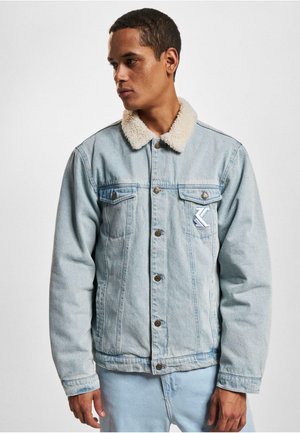 Veste en denim bleu clair avec un col en sherpa beige, fermetures à bouton et deux poches poitrine arborant un patch logo bleu clair.