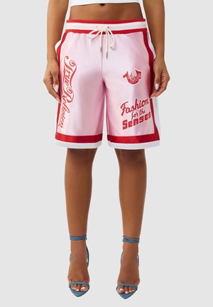 Donna che indossa pantaloncini di raso rosa con bordi rossi e coulisse bianca, con disegni di testo rossi, abbinati a sandali con tacco blu con laccio alla caviglia.