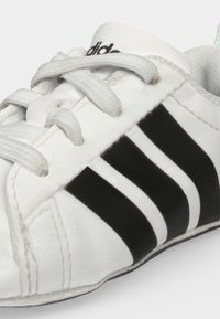 Zapatilla blanca con rayas negras, cordones blancos y costuras visibles, mostrando parte de la lengüeta con un logo negro sobre fondo blanco.