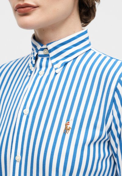 Κοντινό πλάνο ενός ατόμου που φοράει πουκάμισο με κάθετες ρίγες μπλε και λευκές, με κουμπιά και γιακά με κουμπί, και ένα μικρό κεντημένο λογότυπο αλόγου.