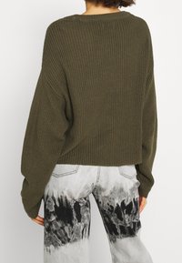 Olivgrüner Strickpullover mit lockerem Sitz und verkürzter Länge, kombiniert mit hellgrauen Jeans aus Denim mit einem schwarz-weißen Batikmuster.