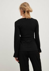 Black long-sleeve top with a textured fabric, fitted design, and a tie at the waist, shown from the back against a neutral background.