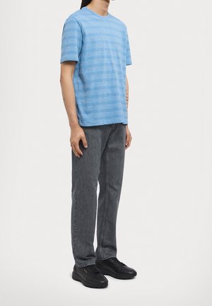 Uomo che indossa una maglietta a maniche corte a righe azzurre, jeans grigio scuro e sneakers casual nere, in piedi davanti a uno sfondo neutro.