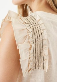 Blusa beige con maniche arricciate e dettagli smock. Tessuto morbido e testurizzato con un leggero scintillio e delicate pieghe lungo i bordi.