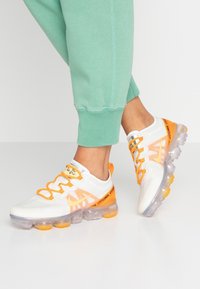 Vita atletisk skor med orange detaljer och mesh-överkänsla, med en transparent sula och snörning, i kombination med gröna joggers.