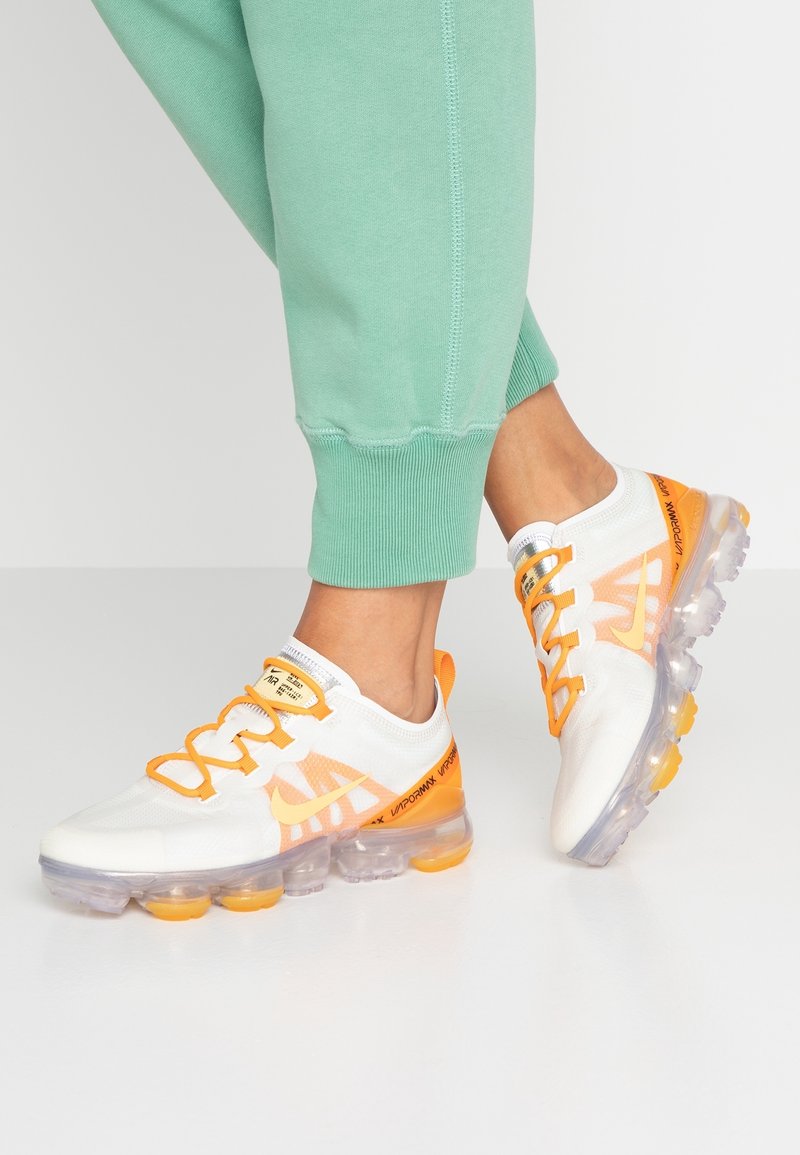 Vita atletisk skor med orange detaljer och mesh-överkänsla, med en transparent sula och snörning, i kombination med gröna joggers.