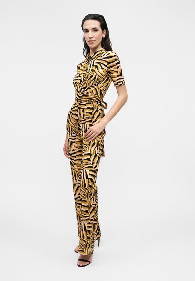 Γυναίκα που φοράει έναν κίτρινο, μαύρο και ροζ ολόσωμο φόρεμα με animal print και κοντά μανίκια, στέκεται με το ένα χέρι στη μέση, λευκό φόντο.