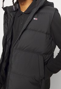 Gilet imbottito nero con design trapuntato, chiusura con zip e tasche laterali, indossato sopra una polo nera. Presenta un piccolo logo ricamato.