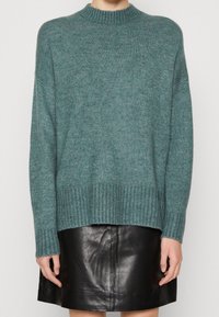 Pull en tricot turquoise avec un col rond côtelé, des épaules tombantes et des manches longues, associé à une jupe en cuir noir.