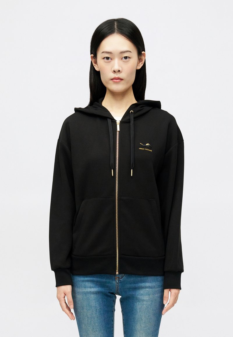 Sweatshirt preto com fecho, feito de tecido suave, apresentando um detalhe em logo dourado no peito, cordões e dois bolsos frontais.