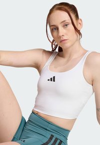 Top corto deportivo blanco con el logo negro de Adidas, textura suave, estilo tank, combinado con shorts de talle alto en color azul verdoso con detalles negros.