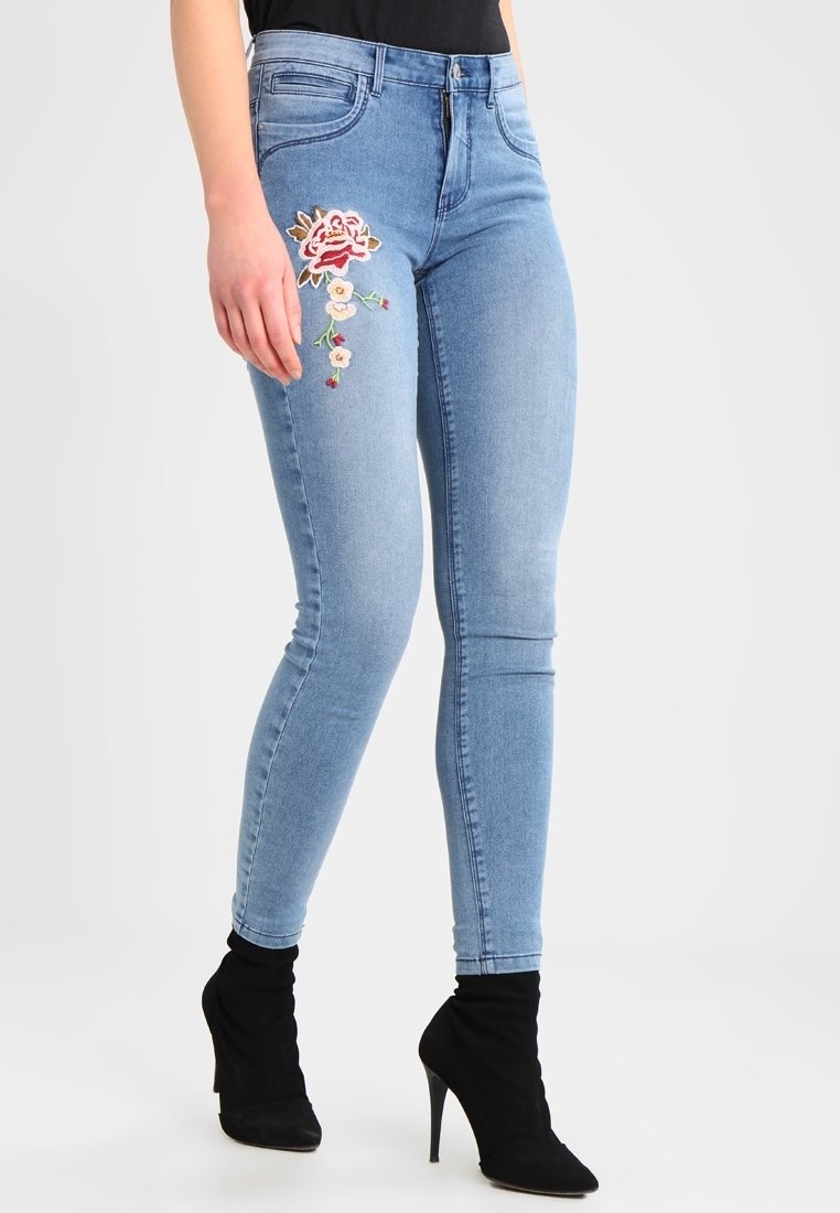Vaqueros pitillo de color azul claro con diseño floral bordado en el muslo izquierdo. Presentan una forma ajustada y un estilo tradicional de cinco bolsillos.