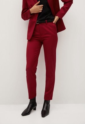 Person trägt eng anliegende dunkelrote Anzughose und passenden Blazer, schwarzes Top, schwarzer Gürtel und schwarze Knöchelstiefel, steht vor einer schlichten Wand.
