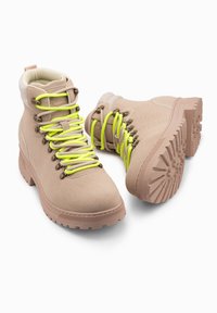 Botas de senderismo de un tono marrón claro con cordones verdes vibrantes, material texturizado similar al ante, collar acolchado y suela de tracción resistente.
