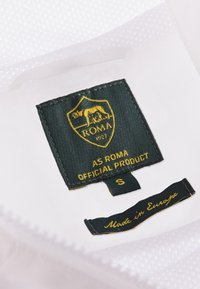 Witte stof met een textuurafwerking bevat een groen label met de tekst "AS ROMA OFFICIAL PRODUCT," met een leeuw-embleem en "Made in Europe." Maat: S.
