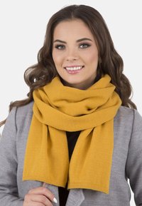 Bufanda de color mostaza hecha de una tela suave y texturizada, drapeada de manera suelta alrededor del cuello. Los bordes son rectos, sin patrones visibles ni herrajes.