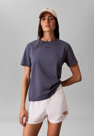 Kobieta w beżowej czapce, ciemnoszarej koszulce Calvin Klein i białych szortach Calvin Klein, stojąca na jednolitym szarym tle.