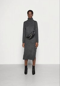 Siva pletena obleka z visokim rolčkom in stranskim razporkom, kombinirana s črno ukrivljeno torbico z verižnim potiskom in obuta v črne gležnjarje.