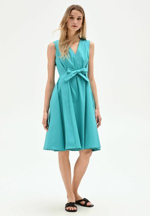 Femme portant une robe turquoise sans manches mi-longue avec un nœud à l'avant, associée à des sandales plates noires et des boucles d'oreilles pendantes dorées.