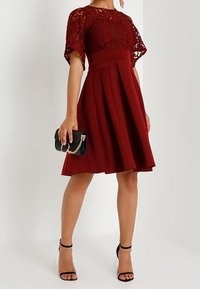 Burgundy klänning med spetstopp, korta ärmar och plisserad kjol. Matchad med svart kuvertväska och remsandaler. Slät textur totalt sett.