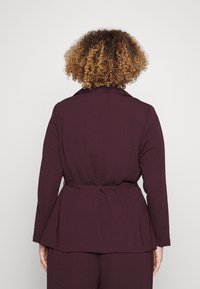 Chaqueta morada de manga larga con una tela texturizada, que presenta un diseño de peplum y una cintura atada. Vista trasera que muestra el cuello y las mangas.