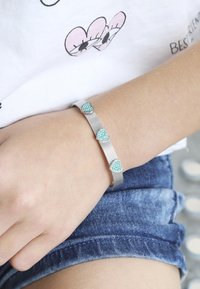Zilveren kettingarmband met drie kleine turquoise hartaccenten, gedragen om een pols boven een dennenstof.