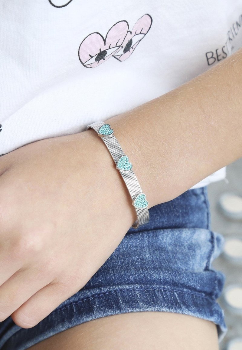 Zilveren kettingarmband met drie kleine turquoise hartaccenten, gedragen om een pols boven een dennenstof.