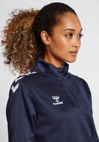 Veste de sport zippée marine avec col haut, arborant un logo blanc et un motif en chevrons sur les épaules, fabriquée dans un matériau lisse et léger.