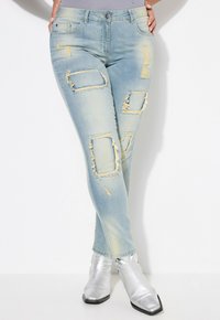 Jean skinny bleu clair avec des détails déchirés, comprenant des rips rectangulaires, des bords effilochés et un léger délavage. Associé à des bottines argentées brillantes.