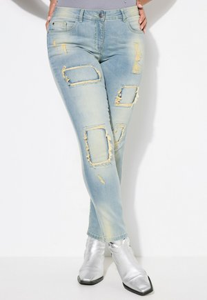 Jean skinny bleu clair avec des détails déchirés, comprenant des rips rectangulaires, des bords effilochés et un léger délavage. Associé à des bottines argentées brillantes.