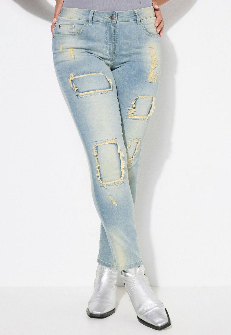 Jean skinny bleu clair avec des détails déchirés, comprenant des rips rectangulaires, des bords effilochés et un léger délavage. Associé à des bottines argentées brillantes.