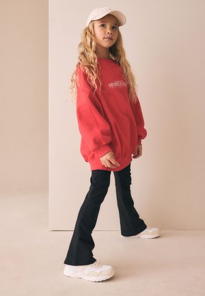 Młoda dziewczyna z długimi blond włosami, ubrana w czerwoną, za dużą bluzę, czarne rozkloszowane spodnie, białe trampki i białą czapkę z daszkiem, stojąca.