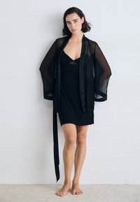 Crni prozirni kimono s širokim rukavima složen preko crne haljine. Haljina sadrži čipkasti detalj i oblik koji se prilagođava tijelu. Mekana tekstura.