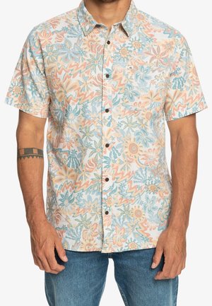 Homme portant une chemise à manches courtes boutonnée avec des motifs floraux pastel et abstraits bleu, orange et beige.