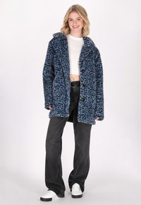 Chaqueta de pelo sintético en azul con estampado de leopardo negro, con un amplio cuello y cierre de botones. Combinada con jeans de pierna ancha negros y zapatillas blancas.